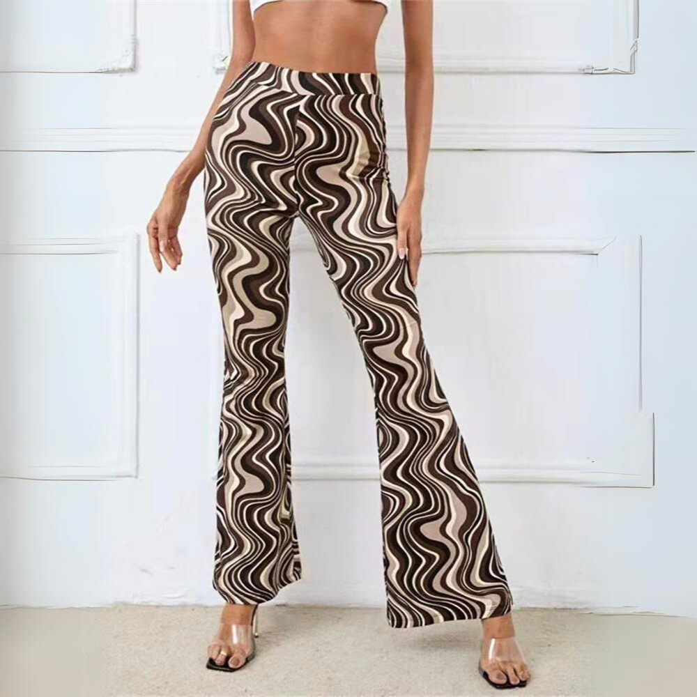 Woman wearing trendy statement pants with a brown, beige, and black wavy swirl pattern in a retro-inspired flare. She pairs the psychedelic flare pants with clear-heeled sandals, photographed indoors against a white wall.
