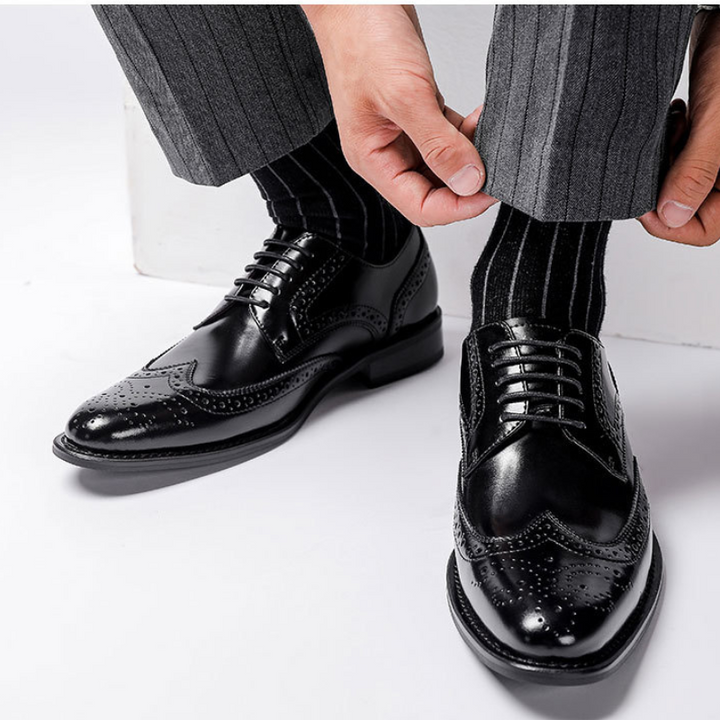 A person adjusts their pinstriped grey trousers while wearing shiny premium leather brogue dress shoes with black socks featuring thin white stripes. The footwear boasts a cushioned insole for added comfort.