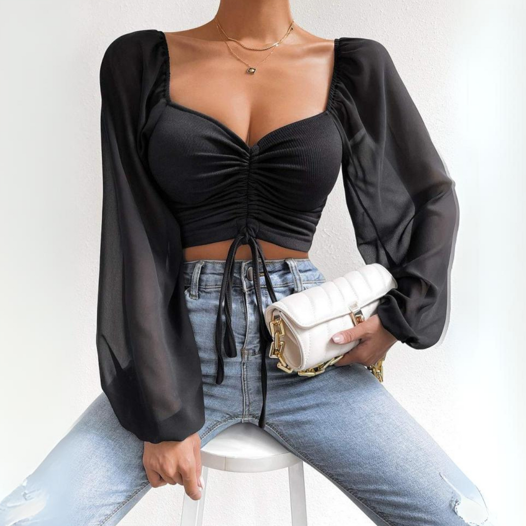 A person wearing a black crop top with a flirty neckline and sheer long sleeves, light blue ripped jeans, a gold necklace, and holding a white handbag, seated against a plain white background.