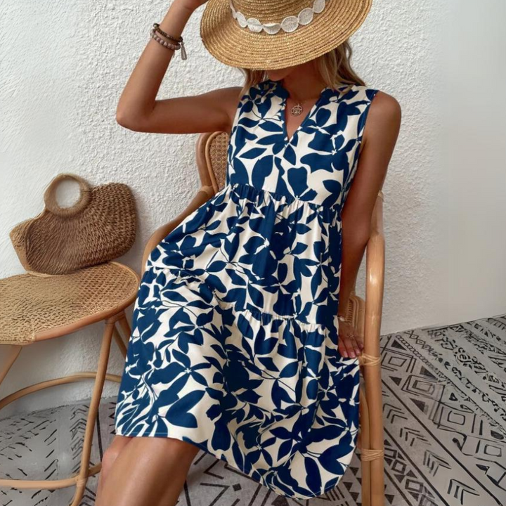 A woman in a sleeveless, blue and white leaf-print dress—crafted from lightweight breathable fabric—sits on a chair. She holds a straw hat, partly covering her face, while a woven bag rests on another chair against patterned flooring.
