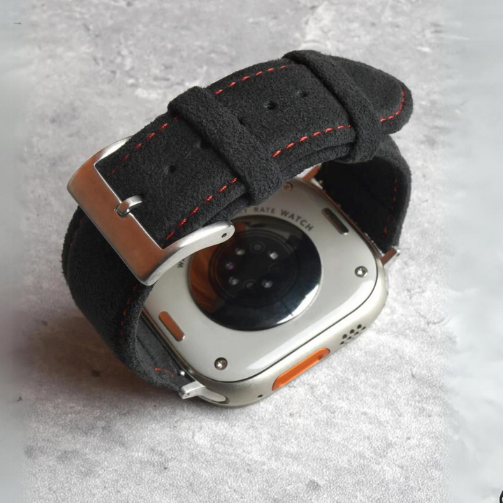 A close-up of the back of a smartwatch with a black Alcantara strap featuring red stitching and a precision fit stainless steel buckle, placed on a textured light grey surface.