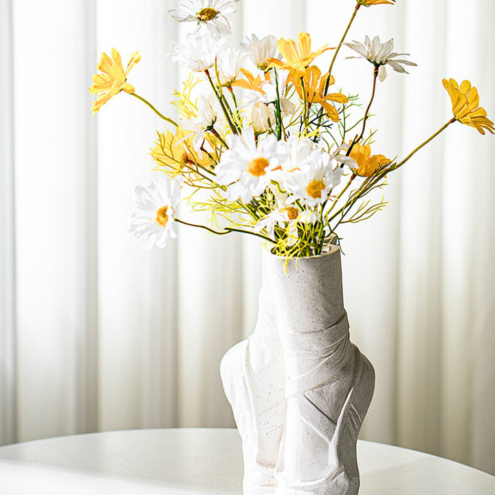 A white ceramic vase with a modern, sculpted design holds a bouquet of daisies, adding contemporary chic décor to a white round table set before sheer curtains.
