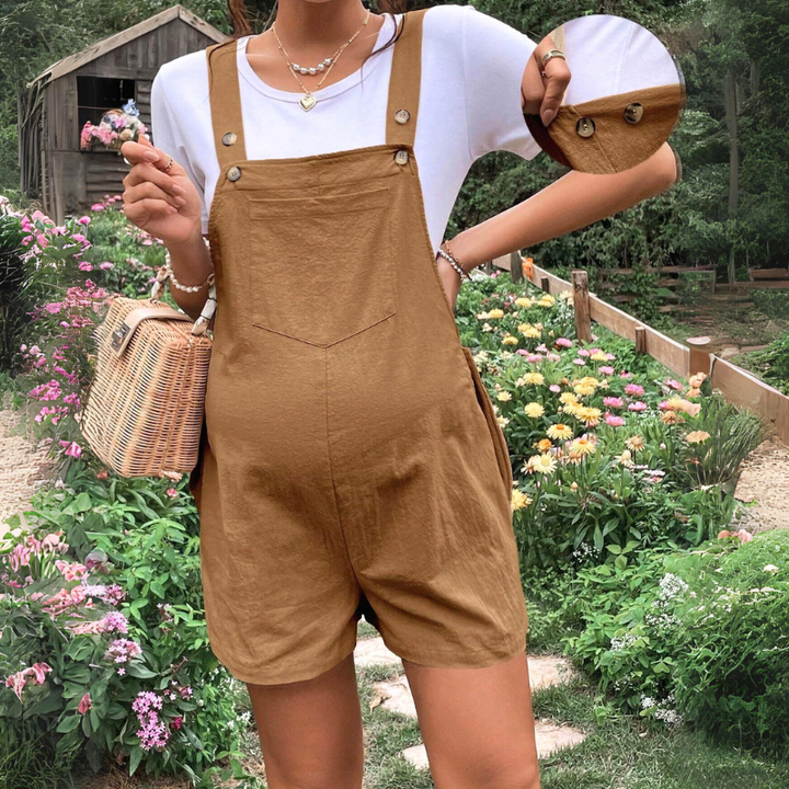 A woman wearing brown pregnancy overalls over a white t-shirt stands in a flower garden, holding a woven bag. The image includes a circular close-up showing button details on the straps, highlighting these comfortable maternity clothes.