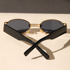 A pair of retro oval sunglasses with a durable metal frame, gold rims, and dark UV400 protection lenses sits on a light surface, with the black arms crossed in an X shape in the foreground.