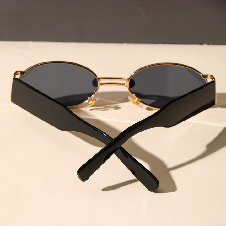 A pair of retro oval sunglasses with a durable metal frame, gold rims, and dark UV400 protection lenses sits on a light surface, with the black arms crossed in an X shape in the foreground.
