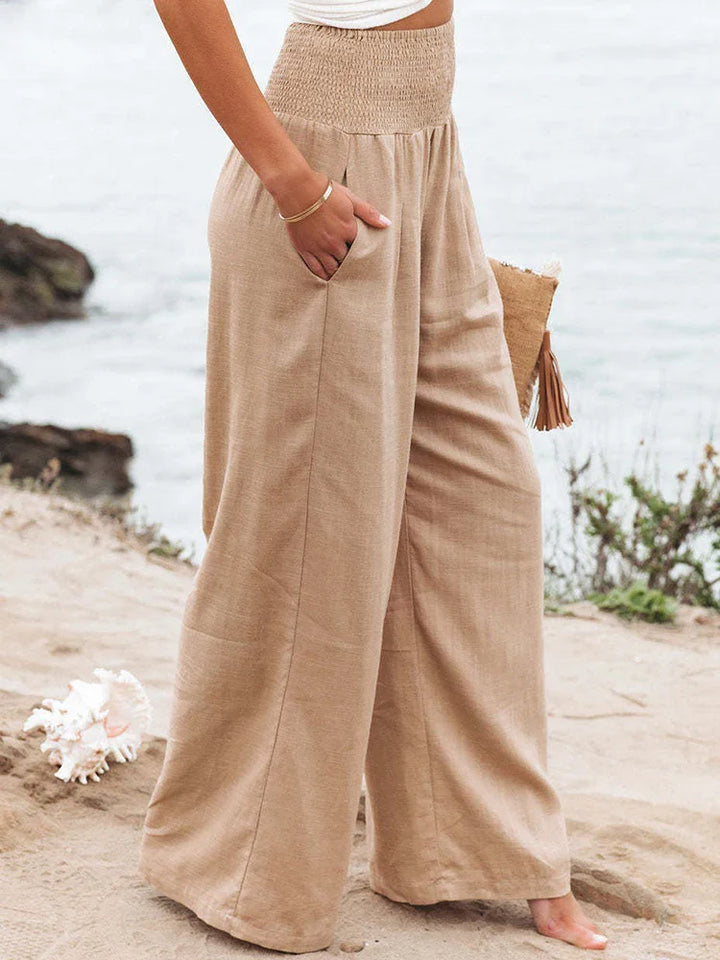 A woman stands on a sandy beach near the water, wearing high waist, wide-leg summer pants and a white crop top, holding a woven clutch with tassels. Some greenery and rocks are visible in the background.