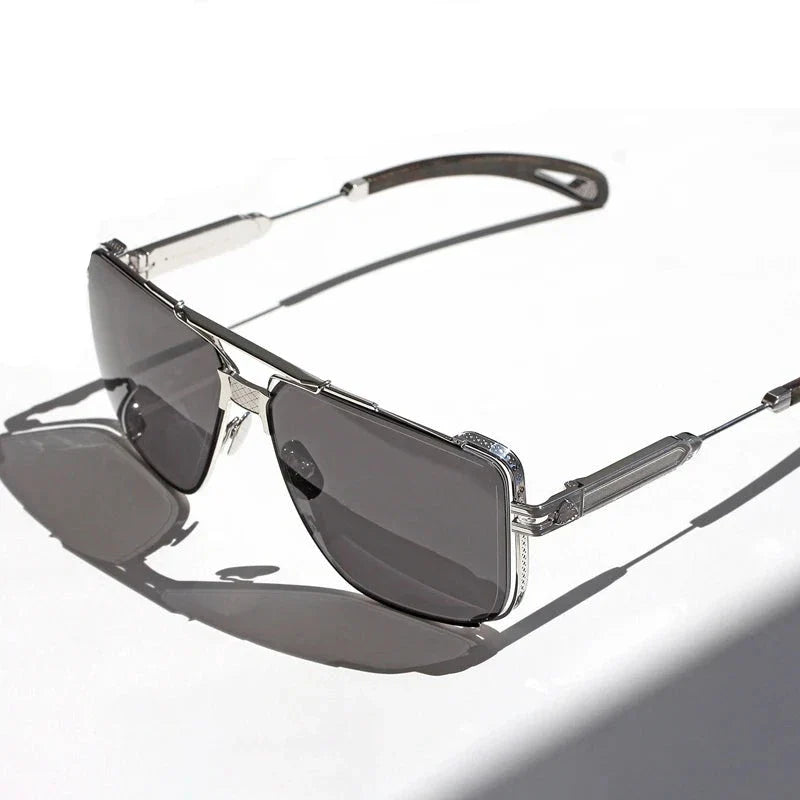 Silver-framed vintage sunglasses with dark tinted, anti-glare lenses rest on a white surface, casting a shadow to the left. The design features intricate detailing on the temples and around the lenses for added style.