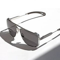 Silver-framed vintage sunglasses with dark tinted, anti-glare lenses rest on a white surface, casting a shadow to the left. The design features intricate detailing on the temples and around the lenses for added style.