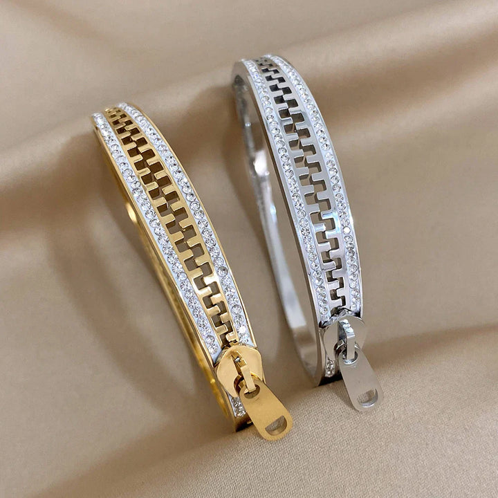 Two metallic zipper bracelets, one a shimmering gold bangle and the other in silver, each adorned with sparkling stones, rest elegantly on a smooth beige fabric background—a striking designer accessory duo.