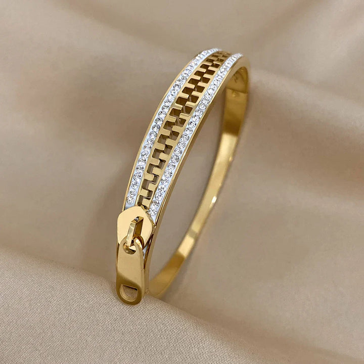 A designer accessory, this gold zipper bracelet features a row of sparkling white stones along both sides, elegantly displayed on a beige fabric background.