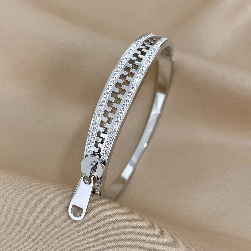 A designer zipper bracelet crafted in silver, with sparkling stones along the edges and a zipper pull charm, is displayed on a beige fabric background—an eye-catching accessory for any jewelry collection.