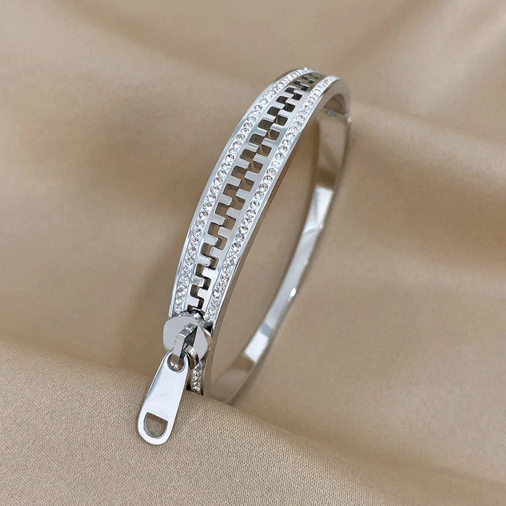 A designer zipper bracelet crafted in silver, with sparkling stones along the edges and a zipper pull charm, is displayed on a beige fabric background—an eye-catching accessory for any jewelry collection.