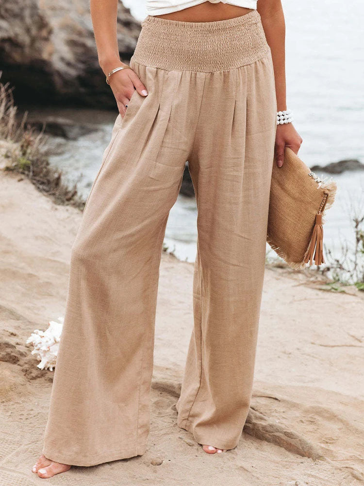 A person stands outdoors by the water, wearing high waist, wide-leg beige summer pants with pleats. They hold a straw clutch with tassels and wear a white beaded bracelet. Only the lower body is visible.