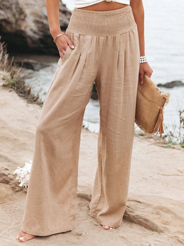 A person stands outdoors by the water, wearing high waist, wide-leg beige summer pants with pleats. They hold a straw clutch with tassels and wear a white beaded bracelet. Only the lower body is visible.
