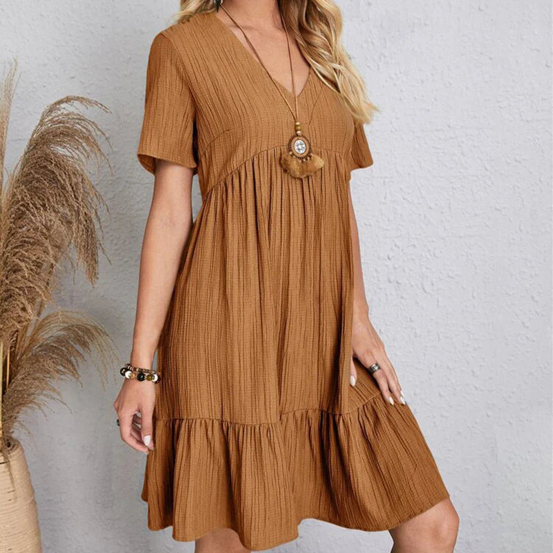A woman wearing a loose-fitting, short-sleeved, knee-length brown dress with a ruffled hem and V-neckline stands against a light gray textured wall. She accessorizes with layered necklaces and bracelets. Pampas grass is visible to the left.
