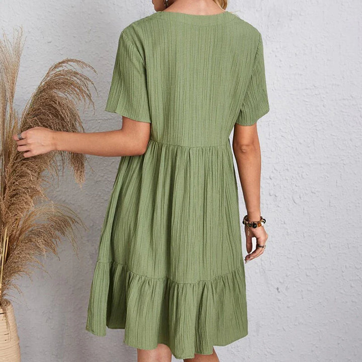 A woman stands facing away, wearing a relaxed, short-sleeved, knee-length green dress with a tiered skirt. She is beside a vase filled with dried pampas grass, set against a light textured wall.