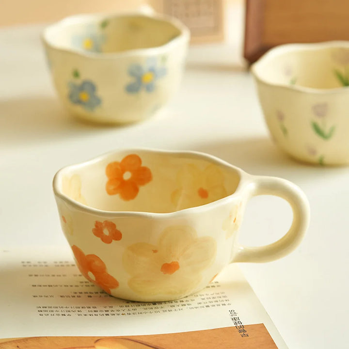 A cream-colored ceramic teacup with orange and yellow flower designs sits on an open magazine. Blurred in the background are two vintage teacups, one with blue flowers and one adorned with small purple tulips.