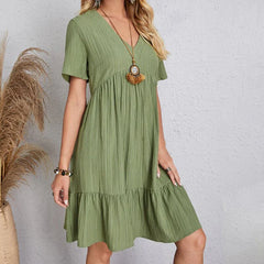 A woman wearing a light green, short-sleeve, V-neck dress with a ruffled hem stands against a light gray wall. She accessorizes with a long necklace and bracelets. Dried pampas grass is elegantly arranged beside her.