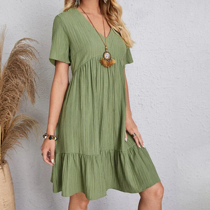 A woman wearing a light green, short-sleeve, V-neck dress with a ruffled hem stands against a light gray wall. She accessorizes with a long necklace and bracelets. Dried pampas grass is elegantly arranged beside her.