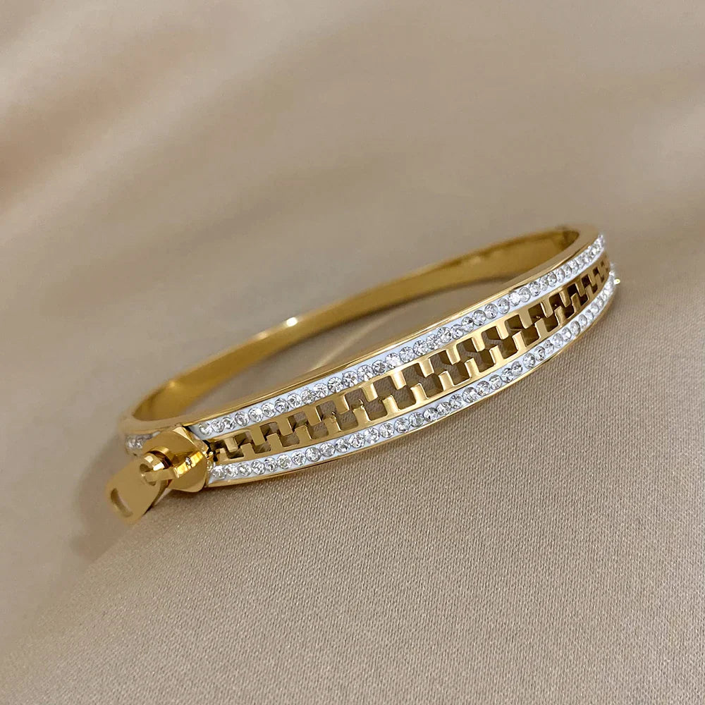 A gold bangle zipper bracelet featuring a row of clear gemstones along both edges and a zipper-inspired design in the center, with a small zipper pull detail on one side, placed on a beige fabric background.