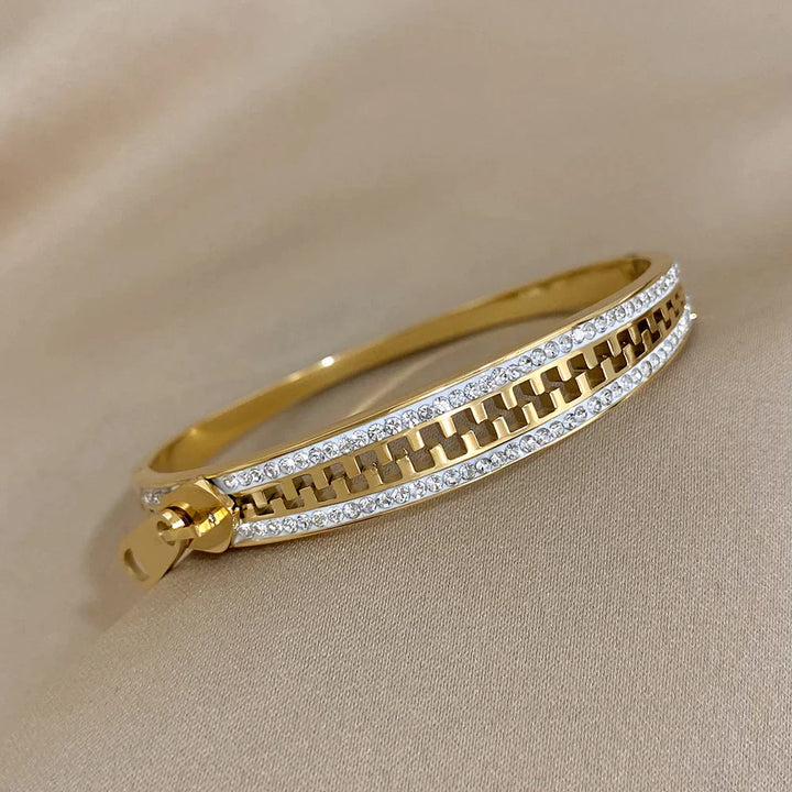 A gold bangle zipper bracelet featuring a row of clear gemstones along both edges and a zipper-inspired design in the center, with a small zipper pull detail on one side, placed on a beige fabric background.