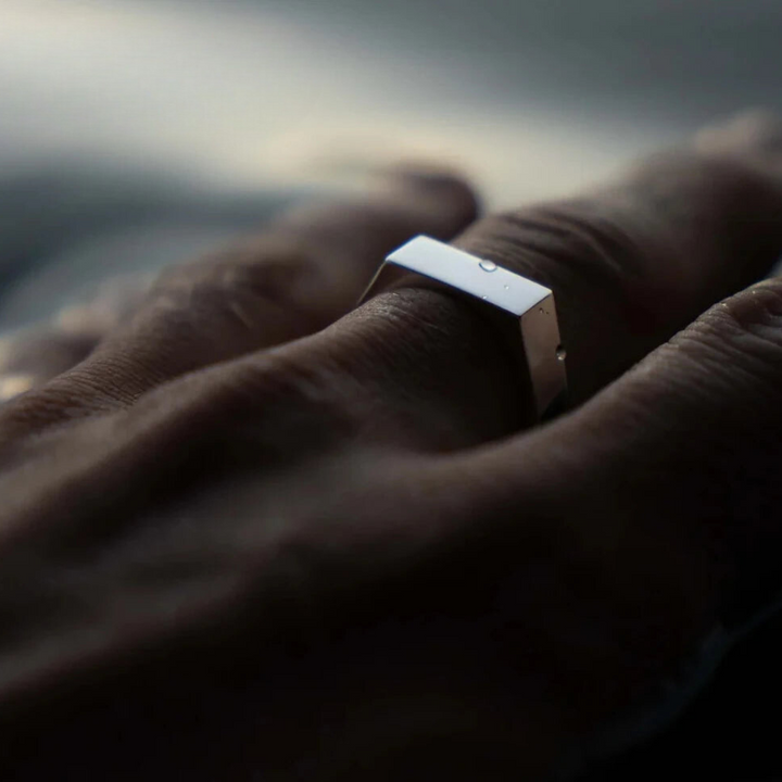 A close-up of a hand wearing a modern, rectangular scratch-resistant silver ring on the middle finger, with soft lighting and a blurred background.