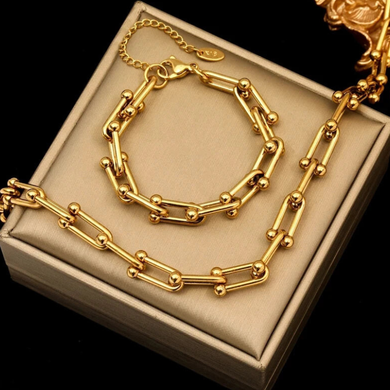 A gold statement chain bracelet and necklace featuring unique bar and bead links with an adjustable clasp are displayed on a beige jewelry box against a black background.