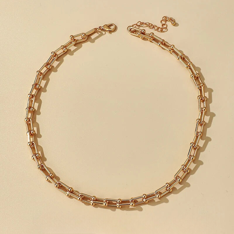 A gold-toned statement chain necklace with rectangular links and small round connectors is laid out in a circular shape on a beige background, crafted from durable 316L stainless steel.