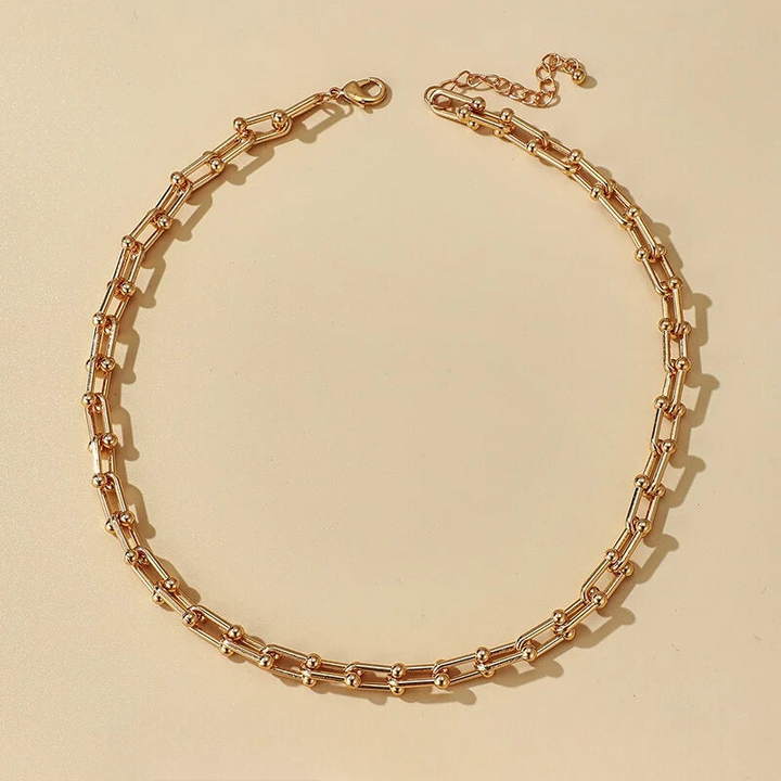 A gold-toned statement chain necklace with rectangular links and small round connectors is laid out in a circular shape on a beige background, crafted from durable 316L stainless steel.