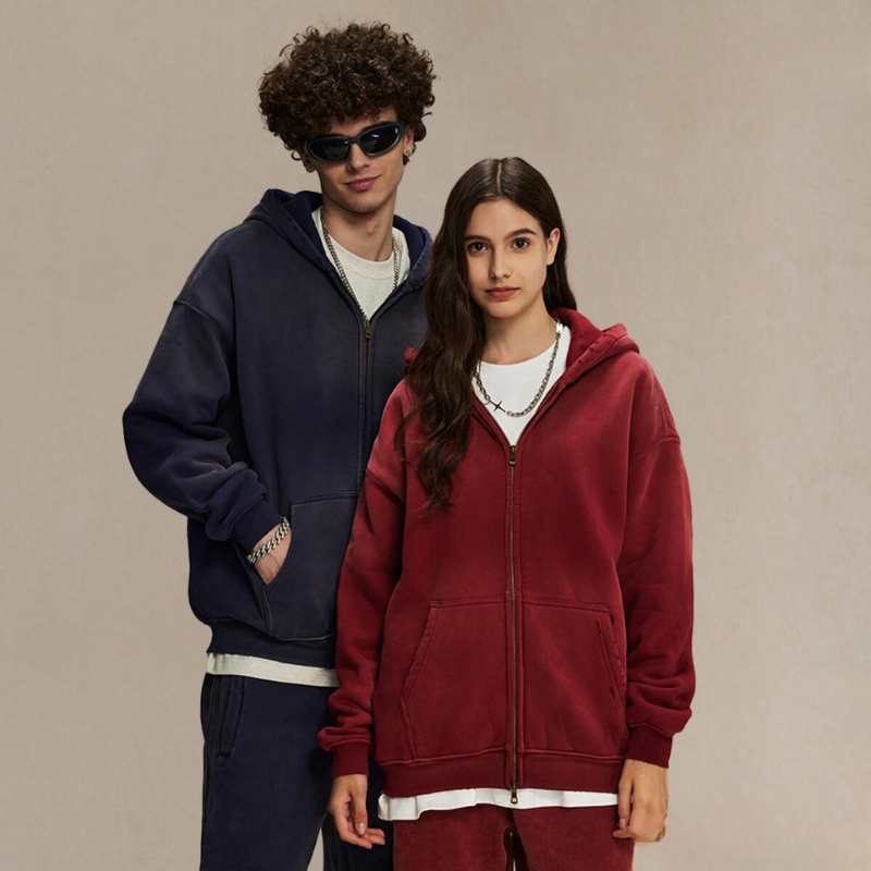 Two people stand side by side wearing relaxed fit, oversized hooded zip-up sweatshirts—one in dark blue, the other in deep red—over light shirts. The person on the left wears sunglasses; both have hands in their pockets.