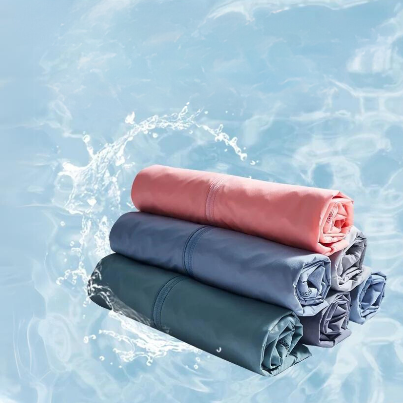 Three neatly rolled towels in pink, blue, and gray are stacked on top of each other against a light blue background with a water splash effect behind them, showcasing their lightweight and quick-dry material.