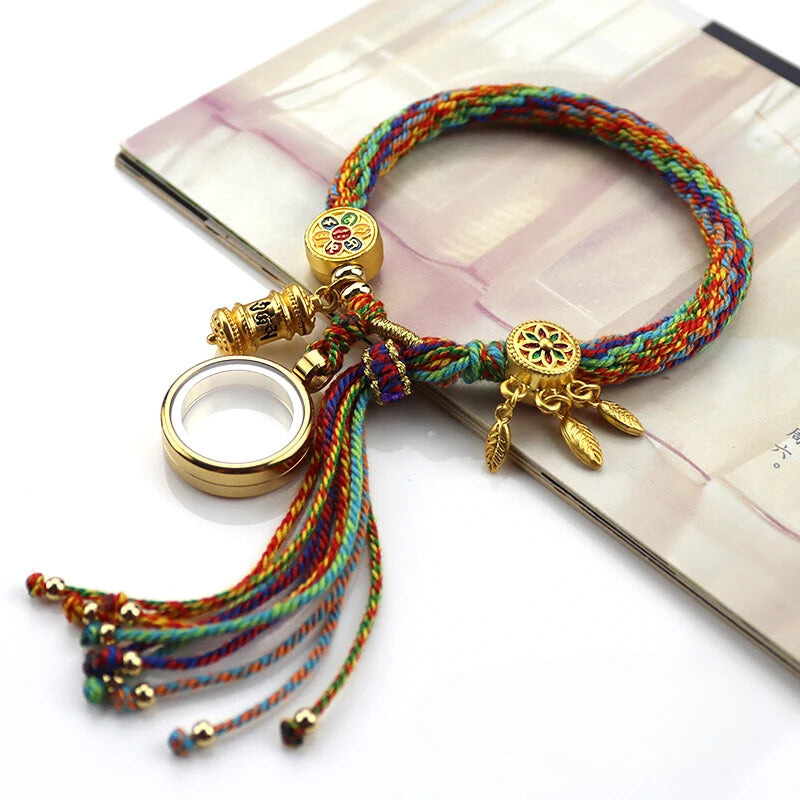 A colorful braided bracelet with gold charms, inspired by Tibetan craftsmanship, including a wheel and small dangling feathers, rests on an open book. Gold beads and a decorative ring accent the adjustable braided strands.
