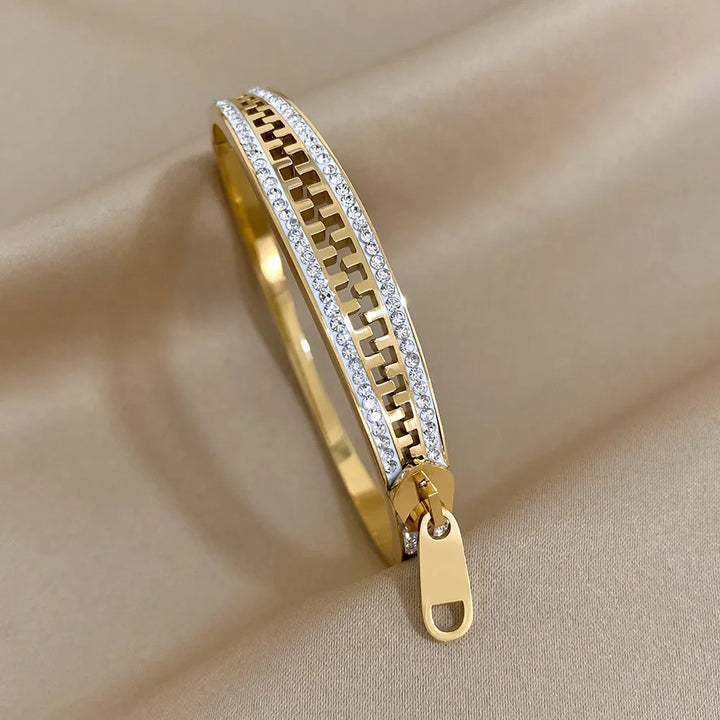 A designer accessory, this gold bangle features a geometric cutout pattern bordered by rows of small white stones and a zipper-like detail at one end, displayed on smooth beige fabric.
