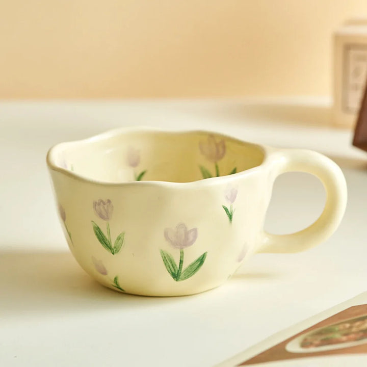 A cream-colored vintage teacup with a wavy rim and handle, decorated with hand-painted light purple tulips and green leaves, sits on a white surface with a soft beige background.