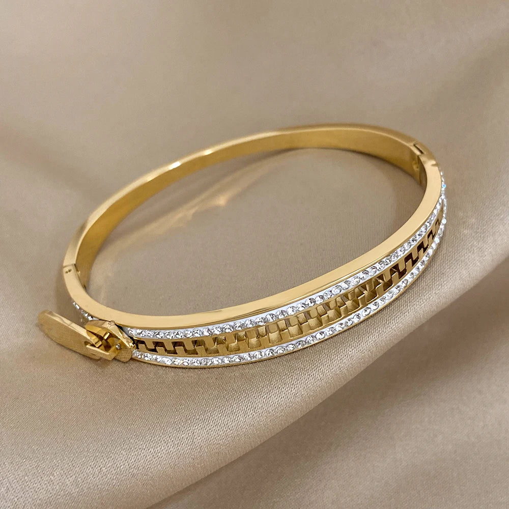 A designer accessory, this gold bangle bracelet features a row of clear, sparkling stones along the edge and a geometric cutout design with a clasp, beautifully displayed on beige satin fabric.