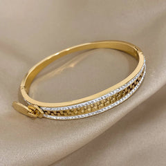A designer accessory, this gold bangle bracelet features a row of clear, sparkling stones along the edge and a geometric cutout design with a clasp, beautifully displayed on beige satin fabric.