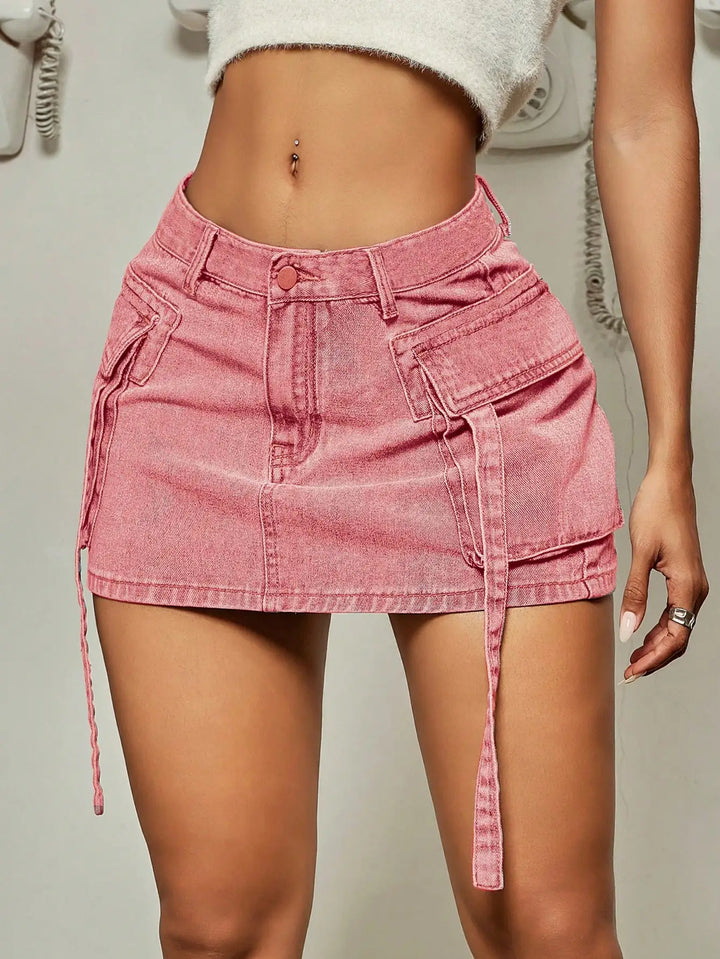A woman wears a high-waisted, pink denim mini skirt with flap pockets, belt loops, side straps, and a frayed hem in a street tooling style, paired with a white crop top that shows her midriff and navel.