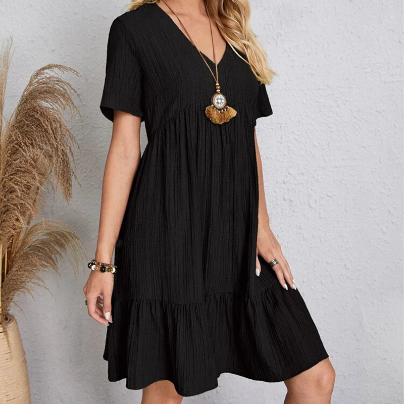 A woman in a short-sleeved, knee-length black dress with a V-neckline stands against a light textured wall. She accessorizes with a long pendant necklace and bracelet, while soft pampas grass adds a touch of nature on the left.