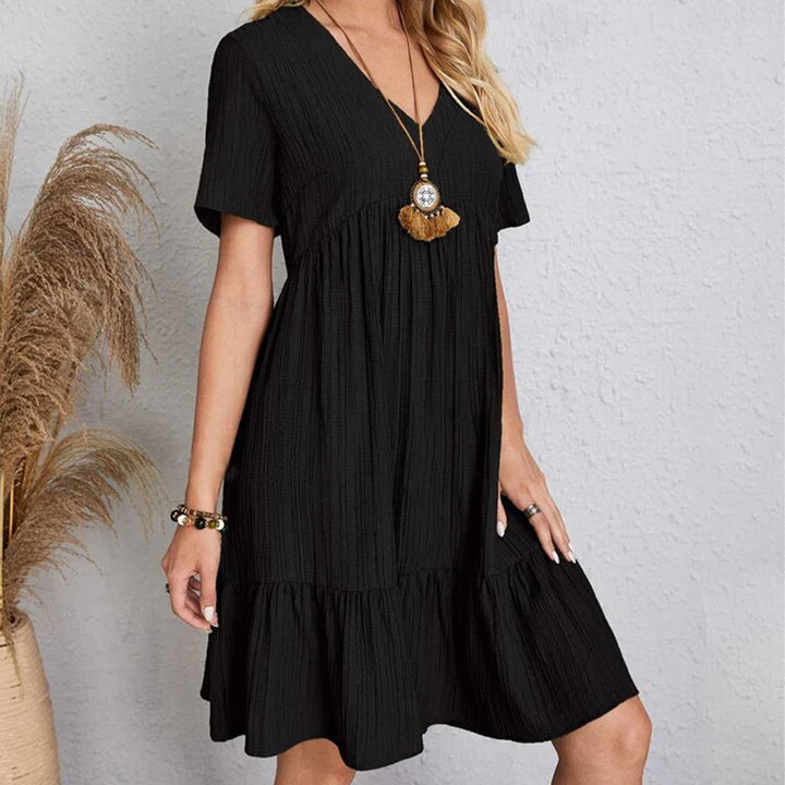 A woman in a short-sleeved, knee-length black dress with a V-neckline stands against a light textured wall. She accessorizes with a long pendant necklace and bracelet, while soft pampas grass adds a touch of nature on the left.