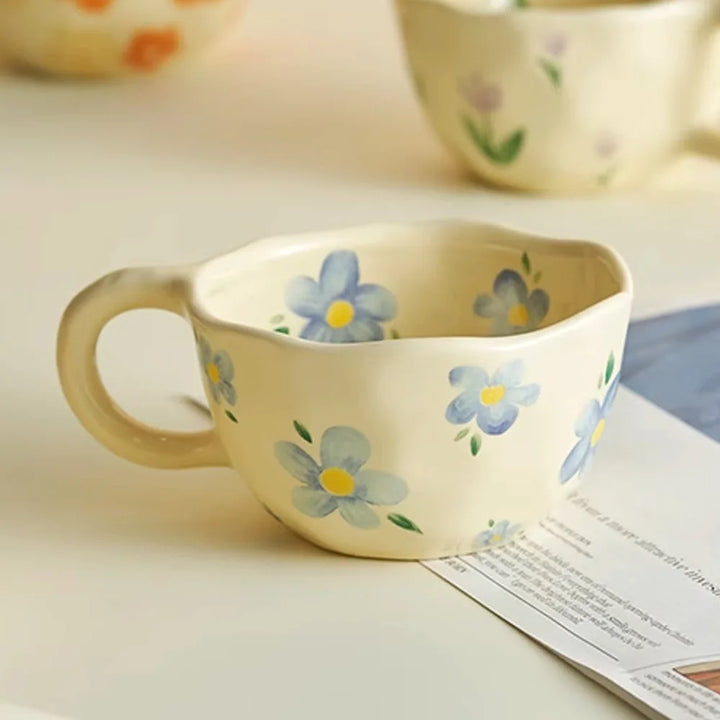 A cream-colored ceramic mug with a round handle, decorated with hand-painted blue flowers and green leaves, sits on a light surface next to some papers. Other vintage teacups with floral designs are blurred in the background.