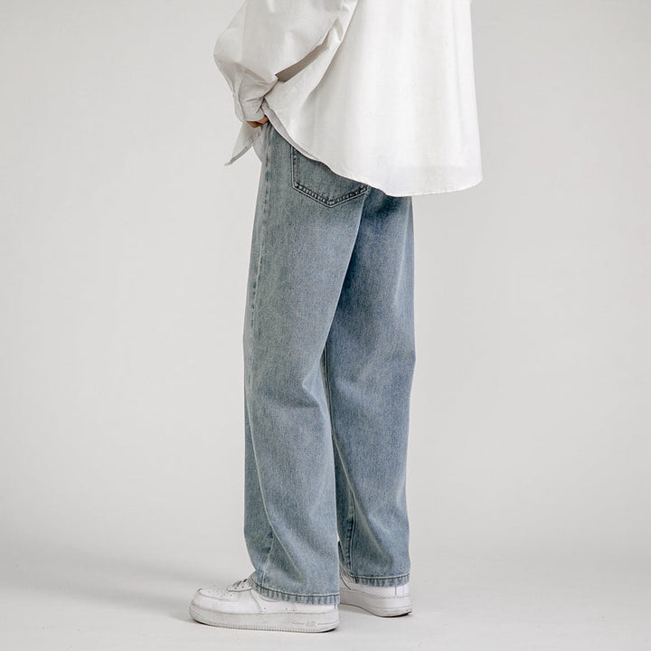 A person wearing a loose white shirt, men’s baggy jeans in a wide-leg design, and white sneakers stands against a plain light background, facing slightly away with hands in pockets for a relaxed Korean fashion vibe.