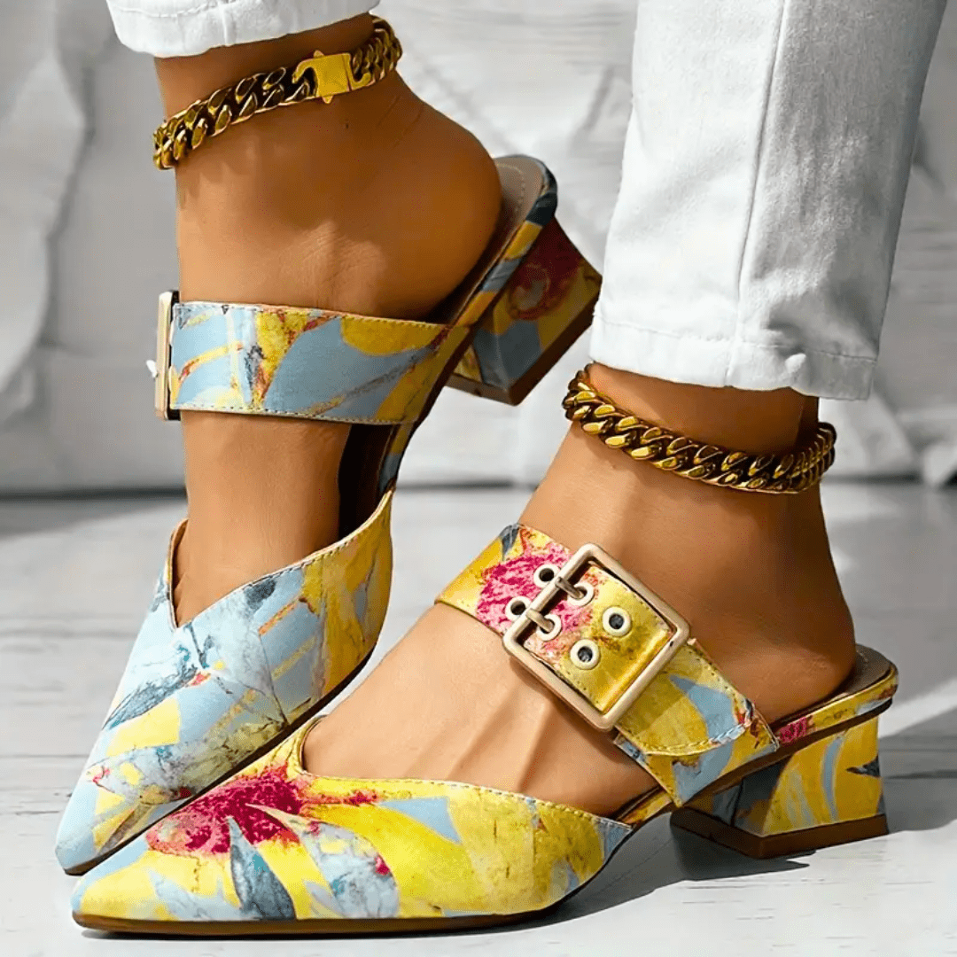 A person wears stylish, colorful floral block heel mules with gold buckle strap and chain anklets. The pointed toe mule shoes feature yellow, blue, and red patterns and pair perfectly with white pants.