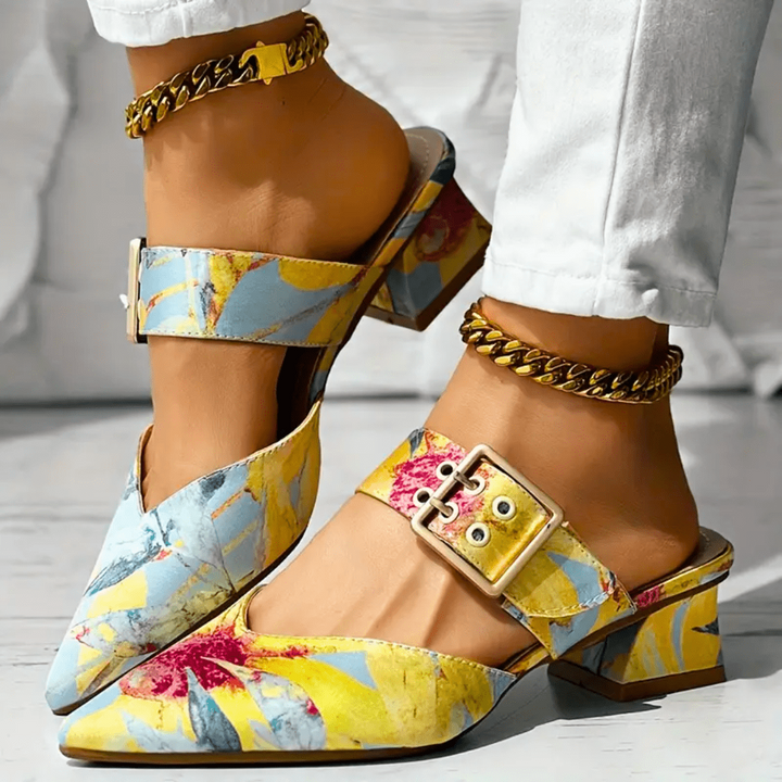 A person wears stylish, colorful floral block heel mules with gold buckle strap and chain anklets. The pointed toe mule shoes feature yellow, blue, and red patterns and pair perfectly with white pants.