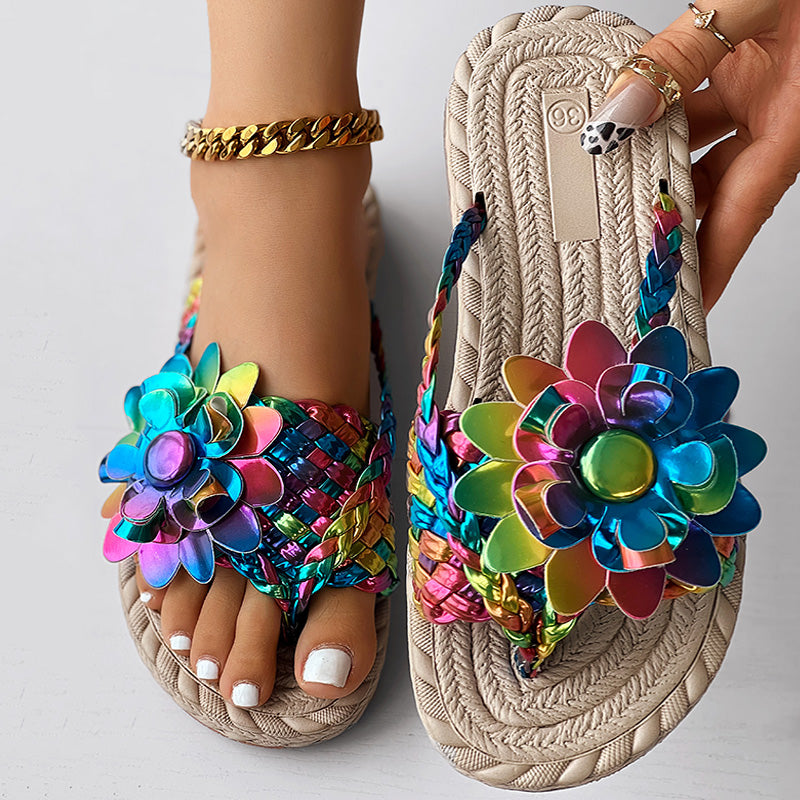 A close-up of a foot in colorful beach flip flops adorned with bold, multicolored flowers. White-painted toenails, gold rings, and an ankle bracelet complete this women's fashion casual shoes look—an ideal vacation essential. Another hand holds the sandal.