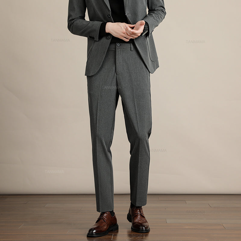 A person wearing a gray suit with Mens Slim Casual Trousers, a black shirt, and brown dress shoes stands on a wooden floor against a beige background. The person’s face is not visible in the image.