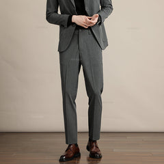 A person wearing a gray suit with Mens Slim Casual Trousers, a black shirt, and brown dress shoes stands on a wooden floor against a beige background. The person’s face is not visible in the image.