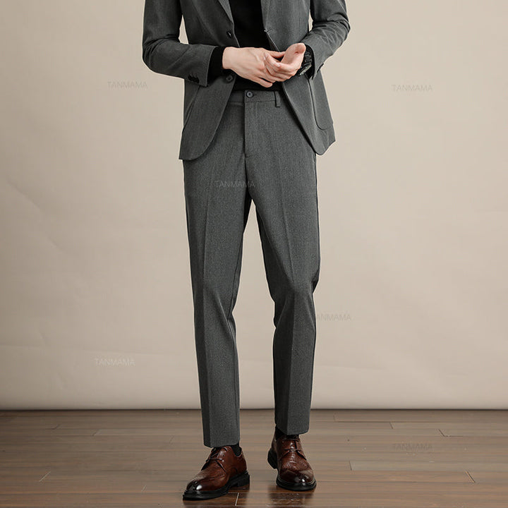 A person wearing a gray suit with Mens Slim Casual Trousers, a black shirt, and brown dress shoes stands on a wooden floor against a beige background. The person’s face is not visible in the image.