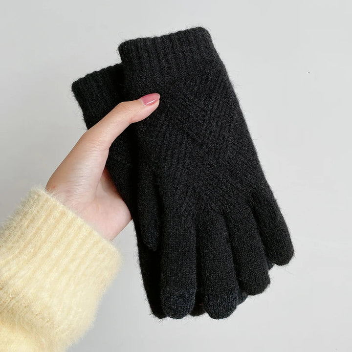A hand with pink-painted nails and a yellow fuzzy sweater holds a pair of luxurious black knit gloves against a plain light gray background.