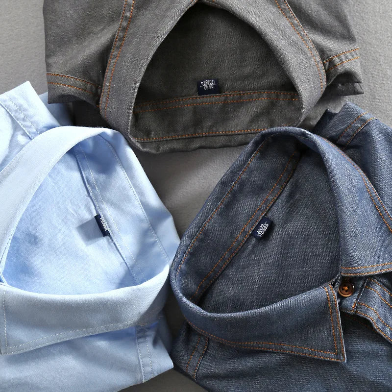 Three neatly folded 100% cotton shirts in gray, light blue, and denim blue, arranged in a circle on a light gray surface. The collars face inward and the shirt tags are visible inside each collar.