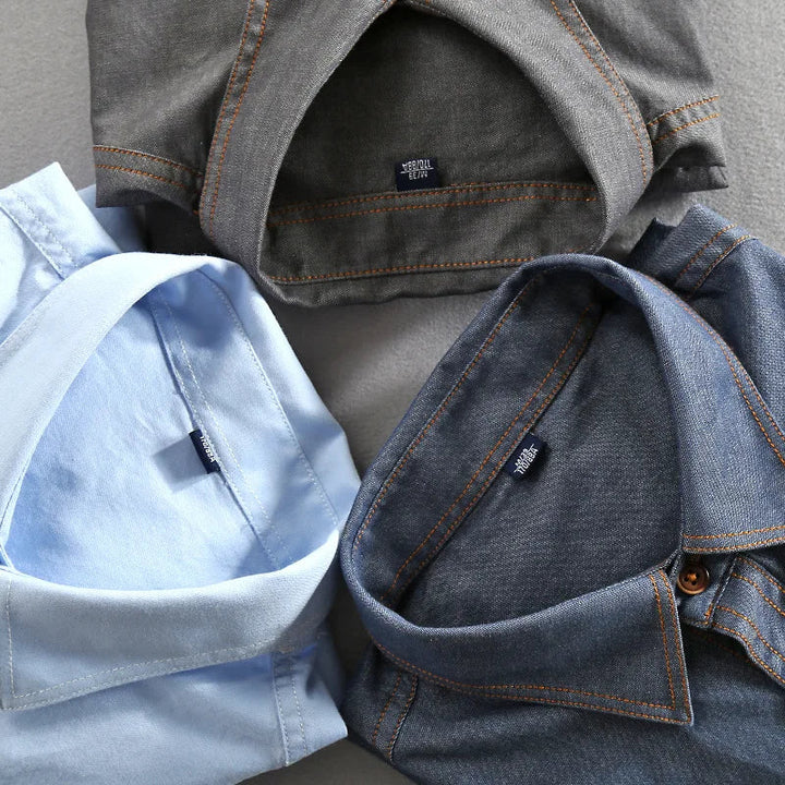 Three neatly folded 100% cotton shirts in gray, light blue, and denim blue, arranged in a circle on a light gray surface. The collars face inward and the shirt tags are visible inside each collar.
